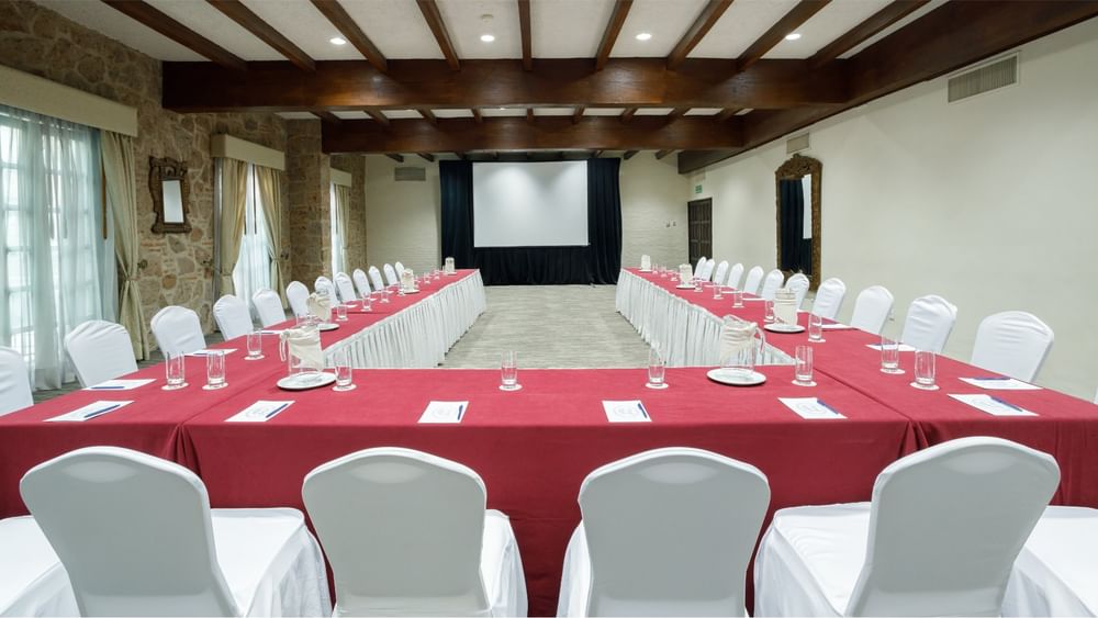 Salón Virreinal with a U-shaped table, red linens, and large screen at Quinta Real Guadalajara