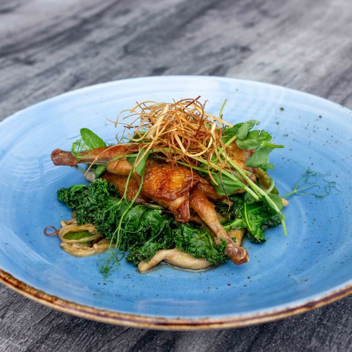 Blue plate with roasted quail, kale, and garnish on a wooden surface.