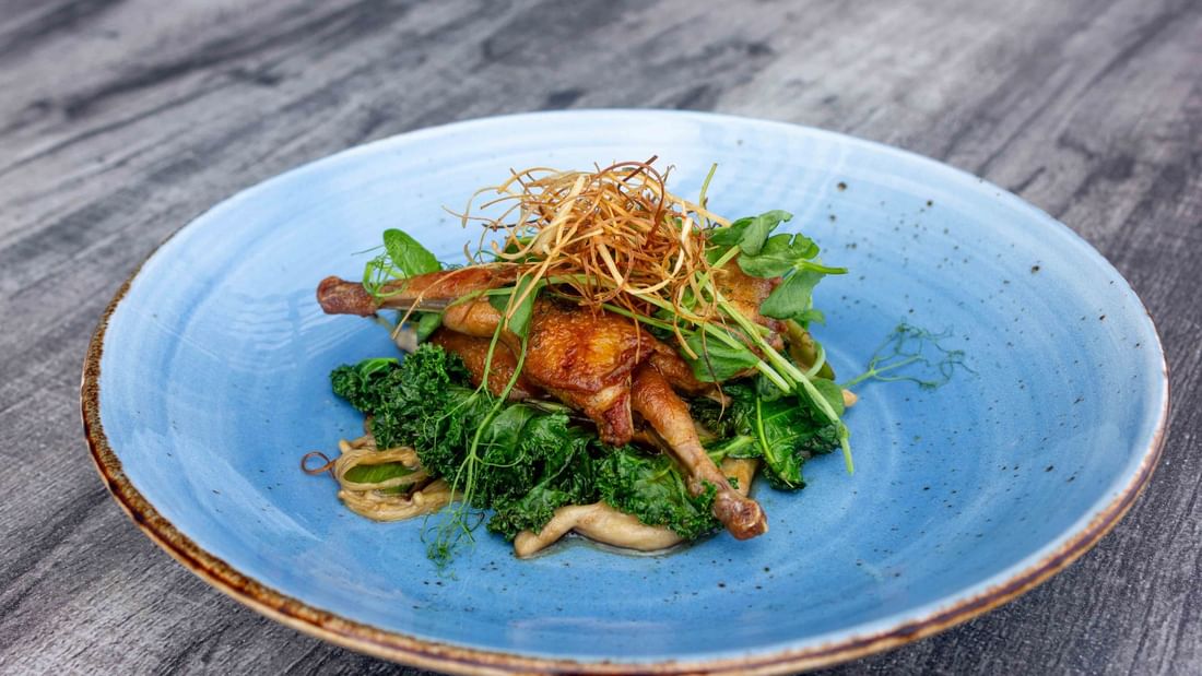 Blue plate with roasted quail, greens, and crispy garnish on a wooden table.