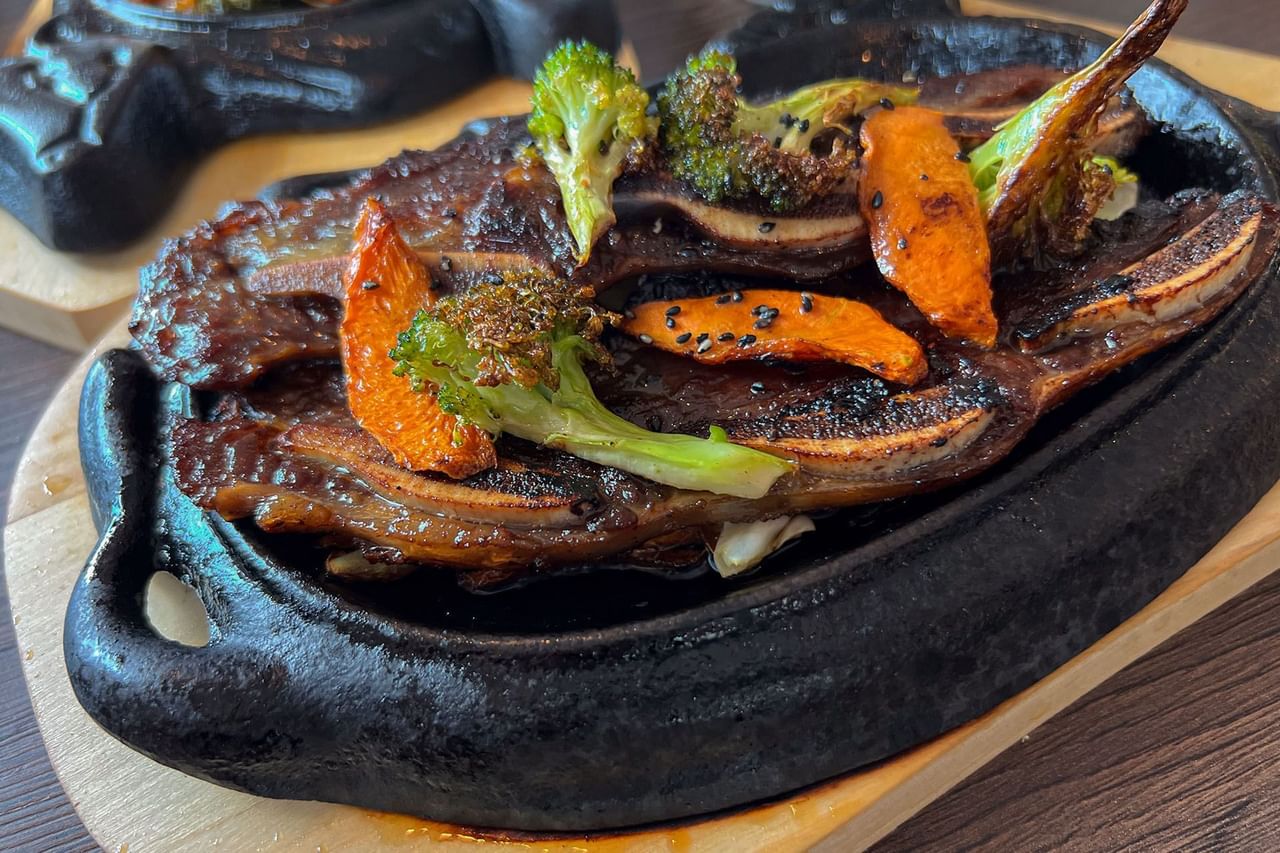A dish of grilled meat with broccoli and carrots on a black plate placed on a wooden surface.