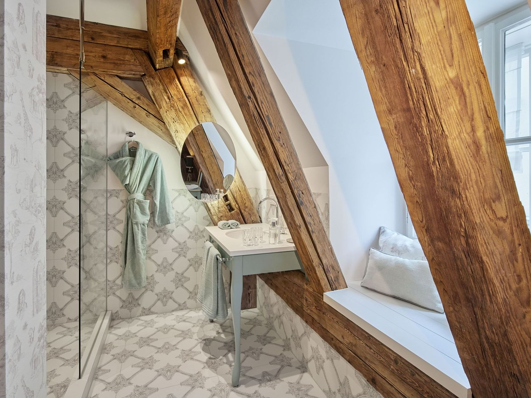 A bathroom with wooden beams, white walls, patterned floor, a sink, mirror, shower stall, and a window with pillows.