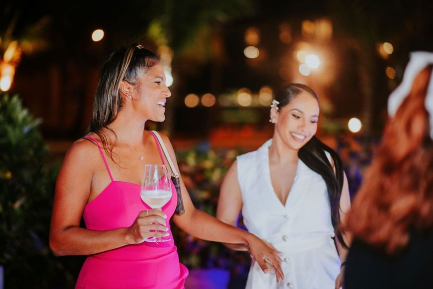 Two women, one in pink and one in white, smiling and holding wine, at an outdoor night event.