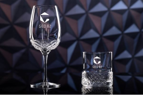 A crystal wine glass and whiskey glass labeled GEO-82 on a sleek black surface with a geometric backdrop.