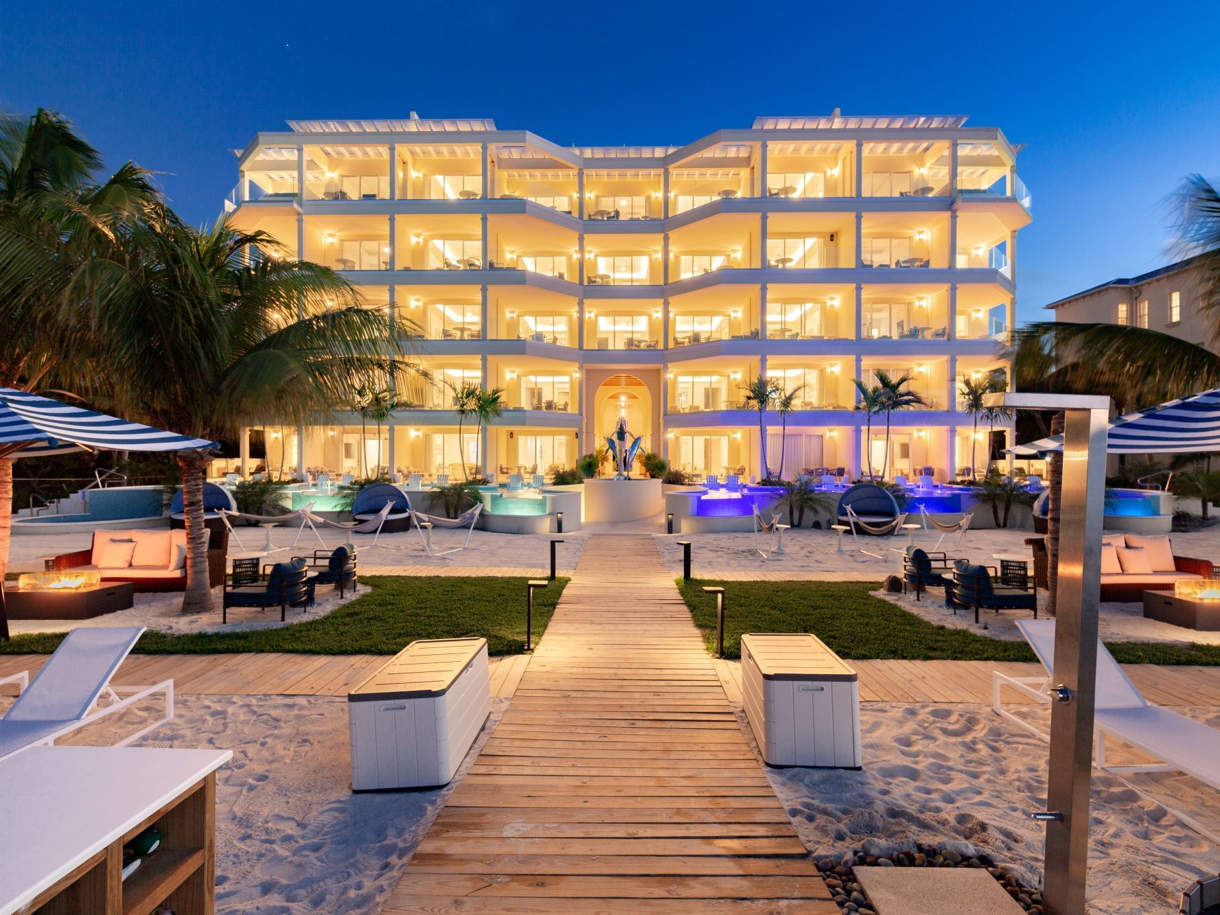 Windsong Resort On The Reef illuminated at dusk, featuring palm trees, a wooden pathway, and inviting spa pools