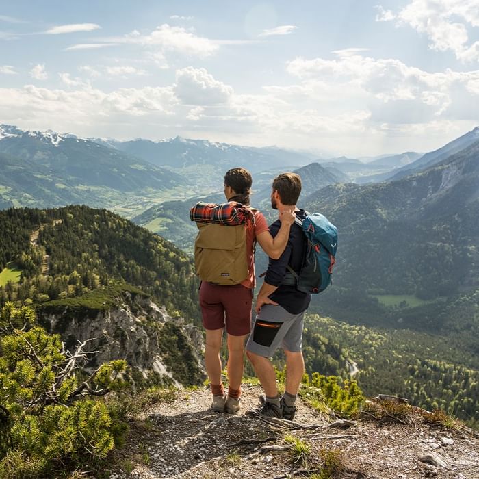 Two hikers with backpacks stand on a mountain peak, admiring the scenic valley and mountain range below.