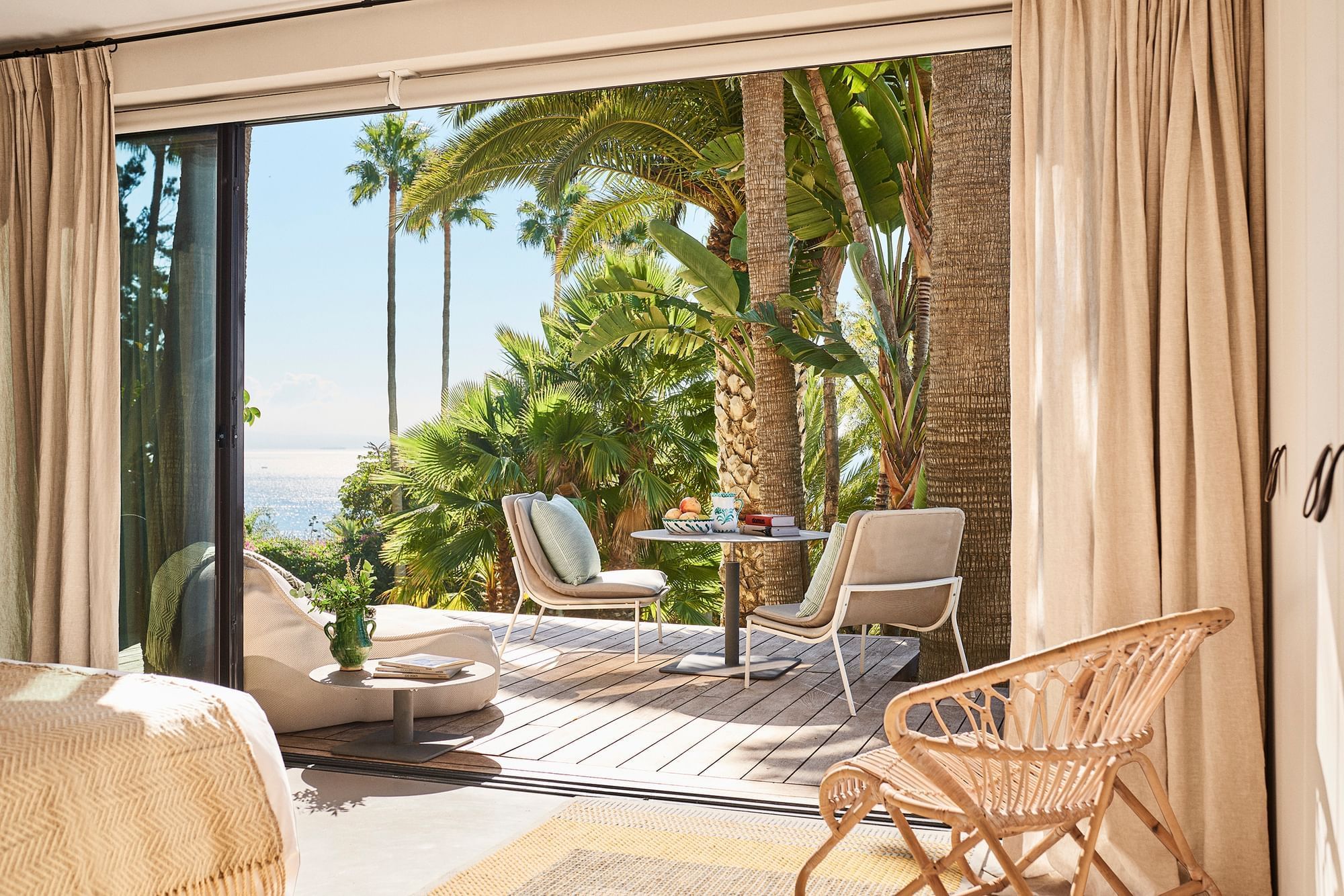 Villa Punta Paloma bedroom overlooking a wooden patio with chairs, swaying palms, and ocean view at Marbella Club resort
