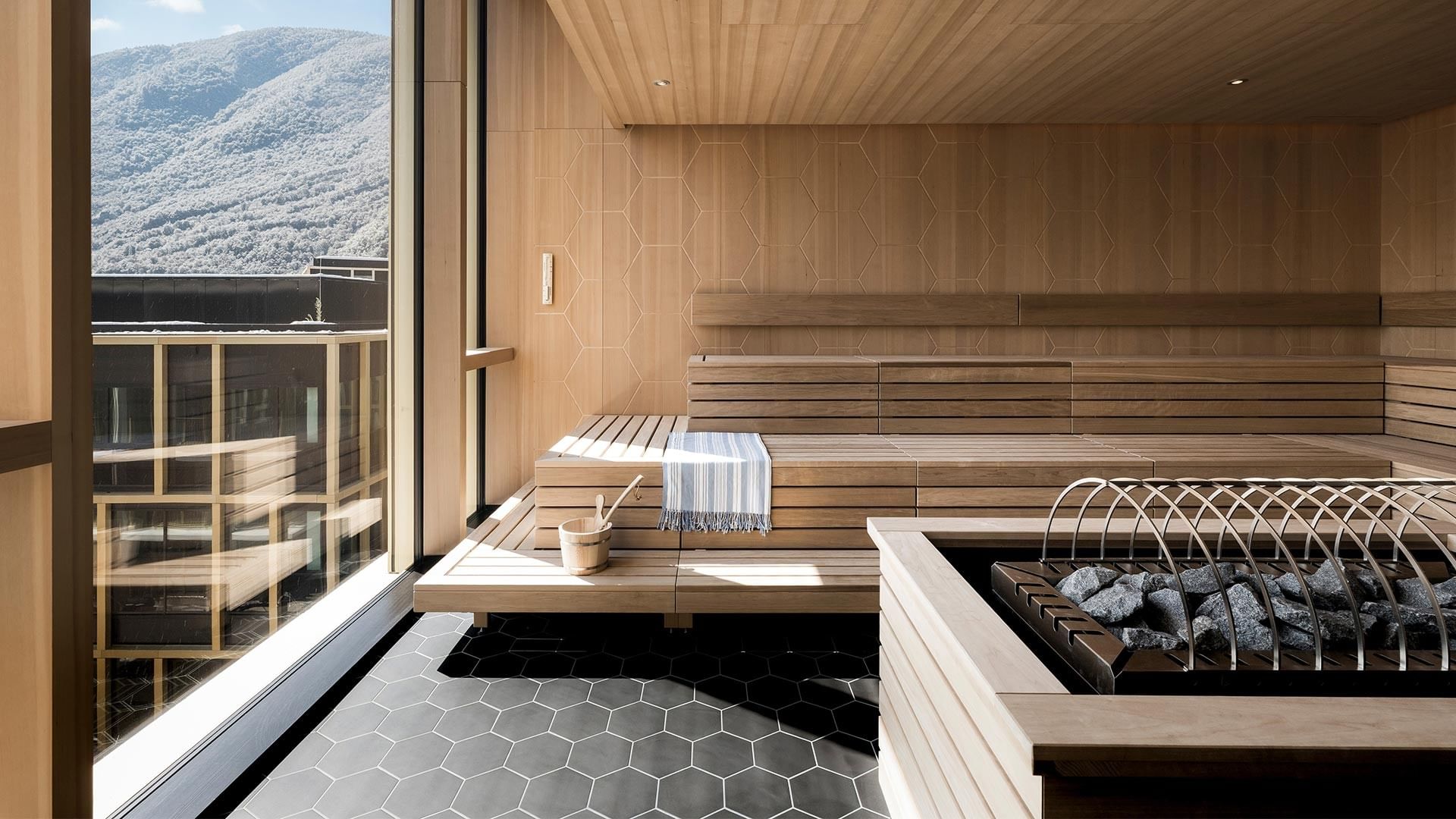 Sauna mit Holzbänken, Steinen und Blick auf Berglandschaft durch große Glaswand.