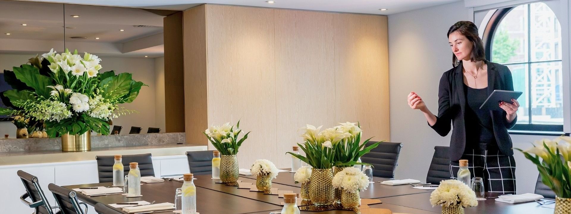 Woman standing at the end of a conference table holding a tablet and speaking to the camera.