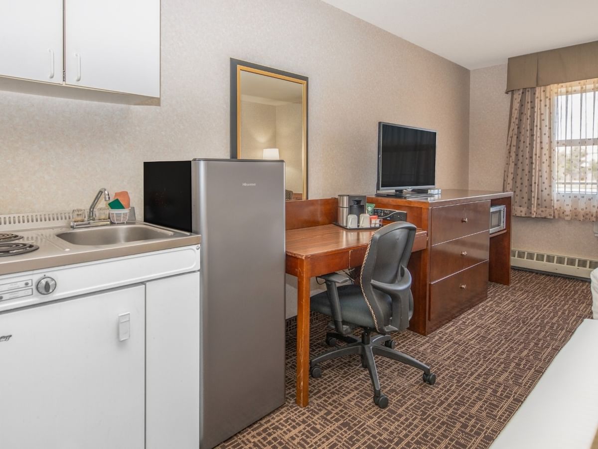 A hotel room with two beds, a small kitchen, a desk with a chair, and a window.