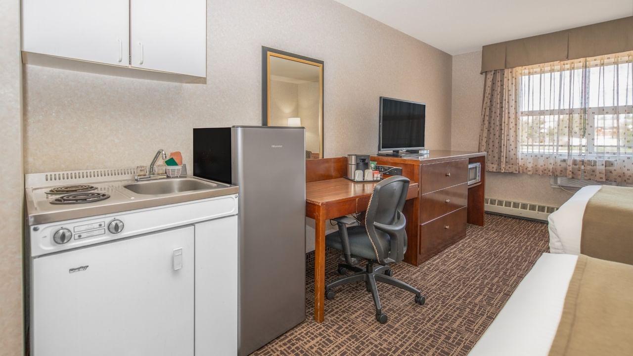 A hotel room with two beds, a small kitchen, a desk with a chair, and a window.