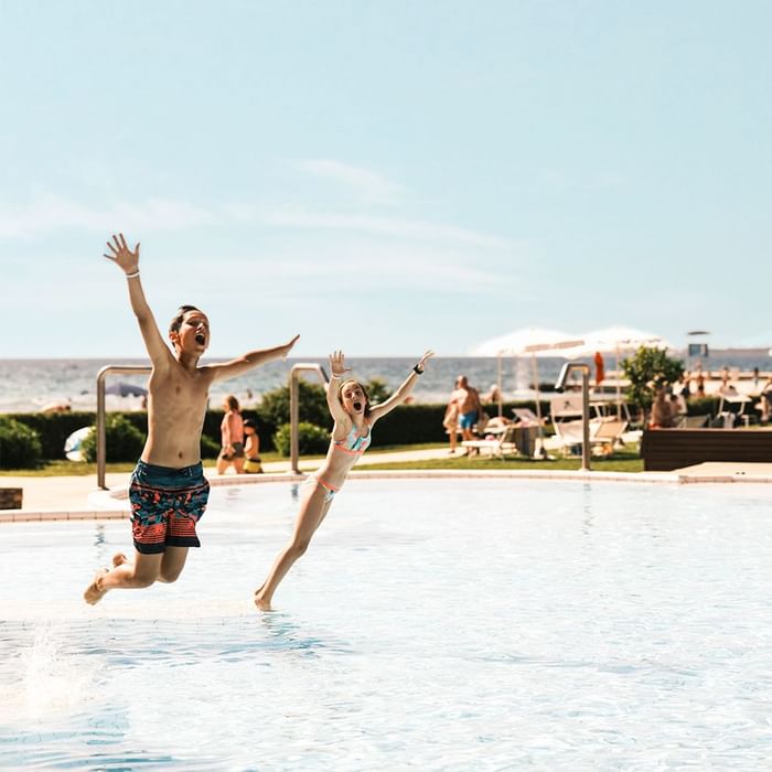 Dječak i djevojčica s rukama podignutim u zrak skakaju u bazen na plaži.