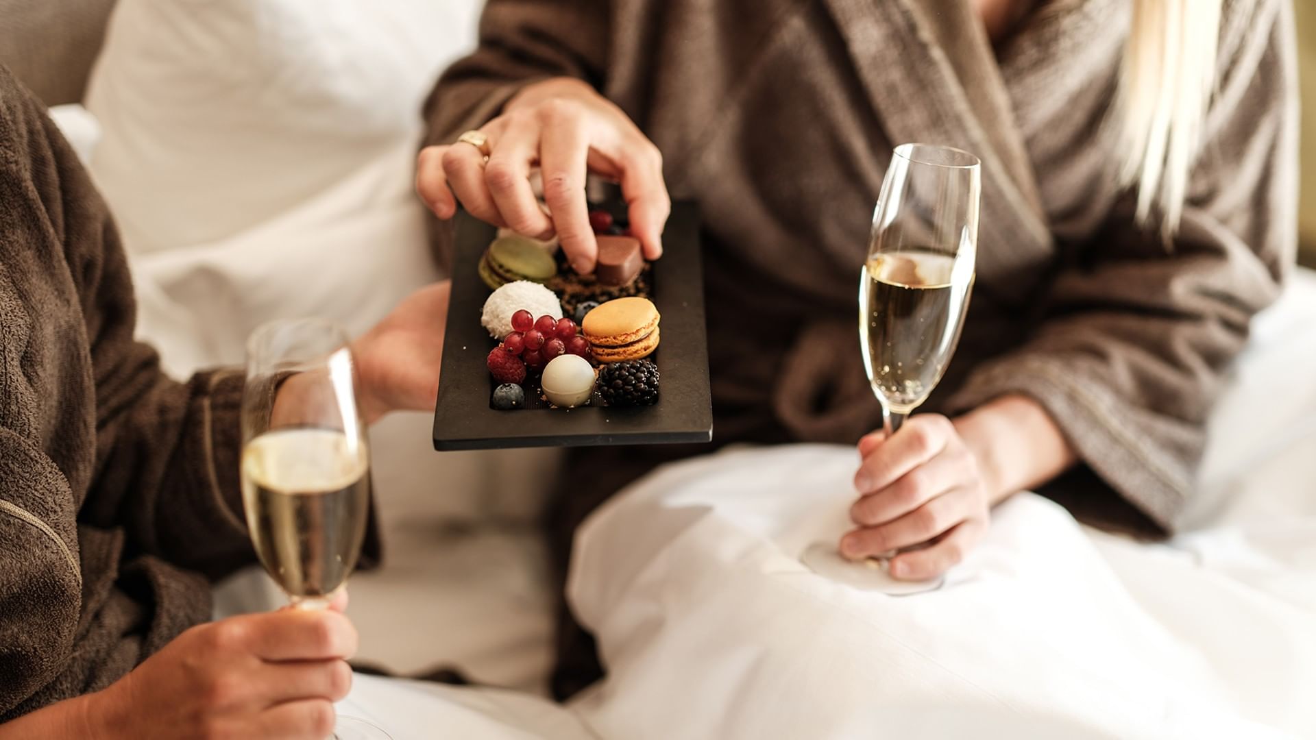 Two people in robes hold champagne glasses while one selects a dessert from a tray.
