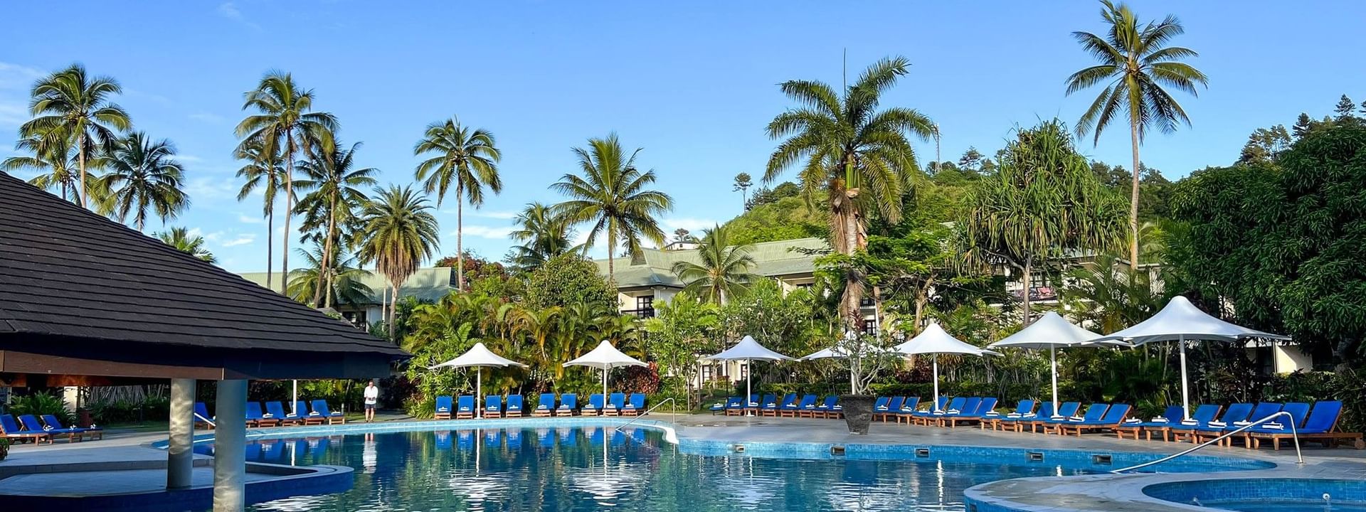 Large swimming pool with blue lounge chairs and umbrellas surrounded by palm trees at The Naviti Resort in Korolevu