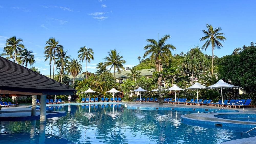 Large swimming pool with blue lounge chairs and umbrellas surrounded by palm trees at The Naviti Resort in Korolevu.