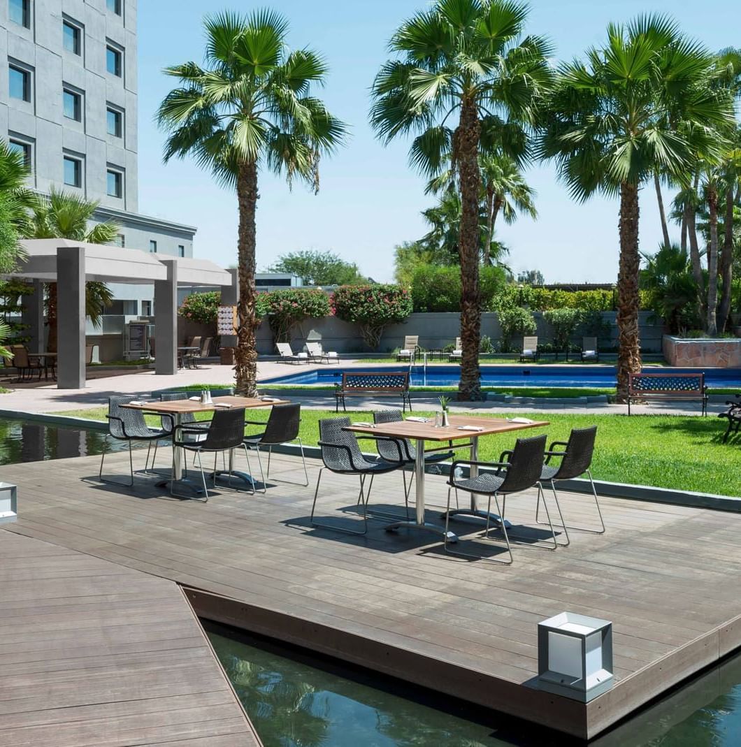 Modern chairs by tables on a wooden deck surrounding a calm pond under palm trees at Real Inn Mexicali