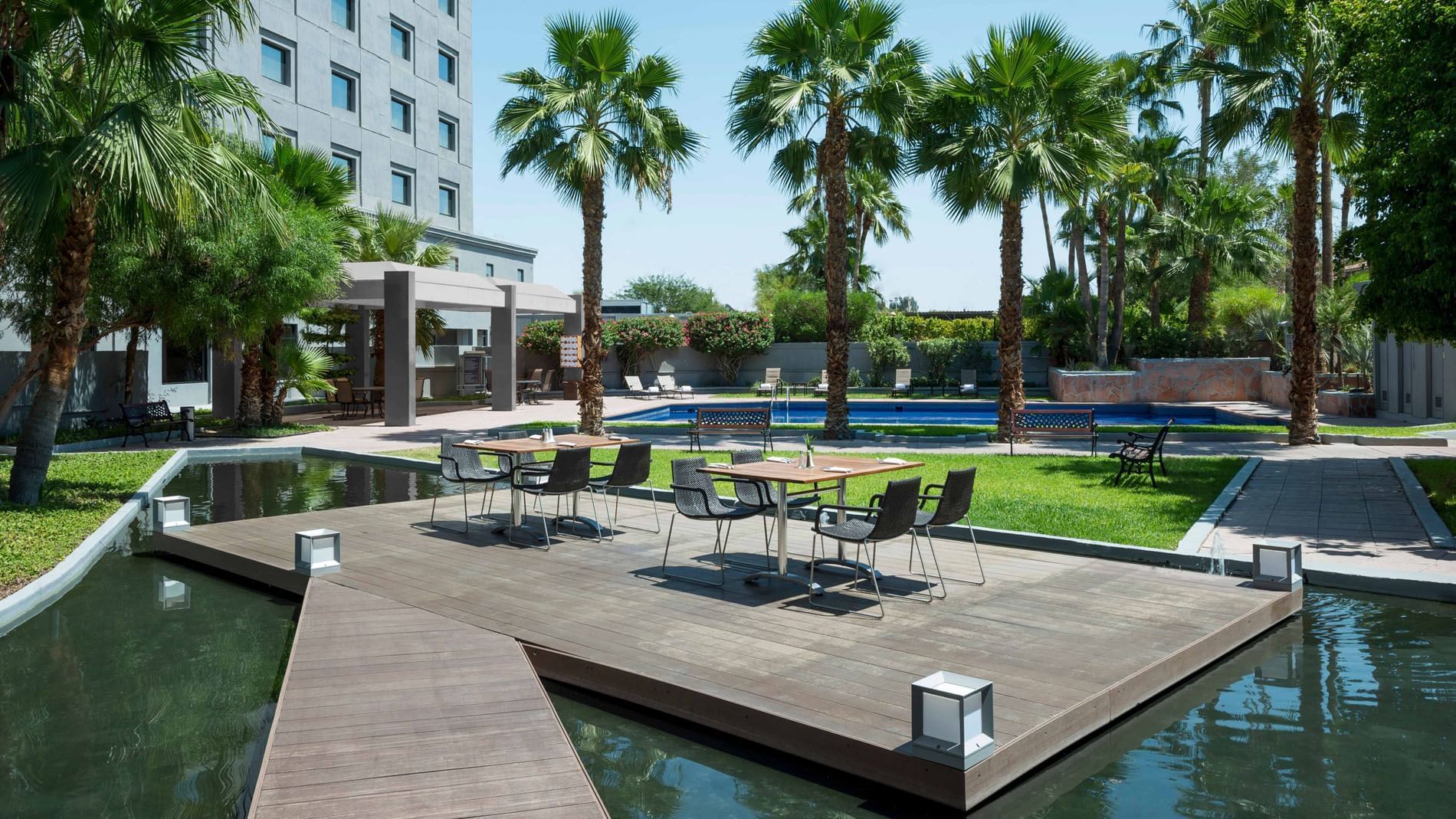 Modern chairs by tables on a wooden deck surrounding a calm pond under palm trees at Real Inn Mexicali