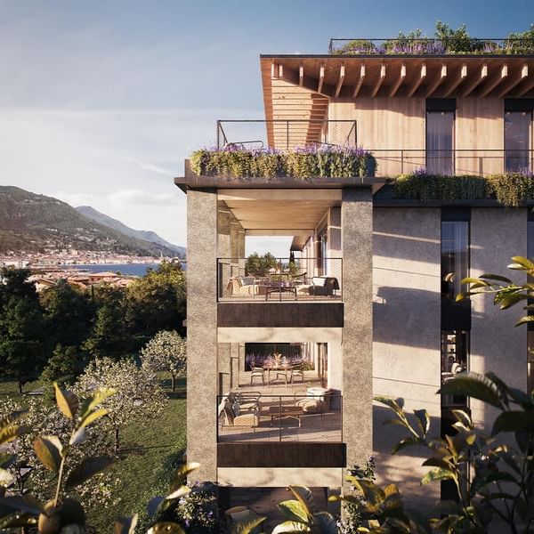 Modern building with balconies and green terraces, surrounded by greenery and mountains in the background.