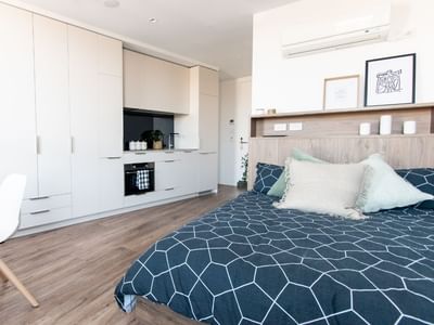 A modern, compact bedroom with a bed, desk, white cabinets, and wooden floors.