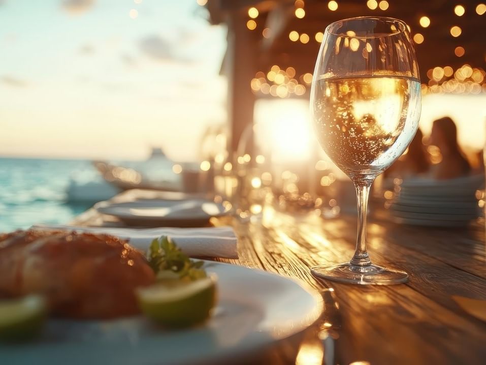 Primer plano de copa de vino y plato de comida sobre mesa de madera con luces bokeh en Fiesta Americana Funeeq Punta Cana