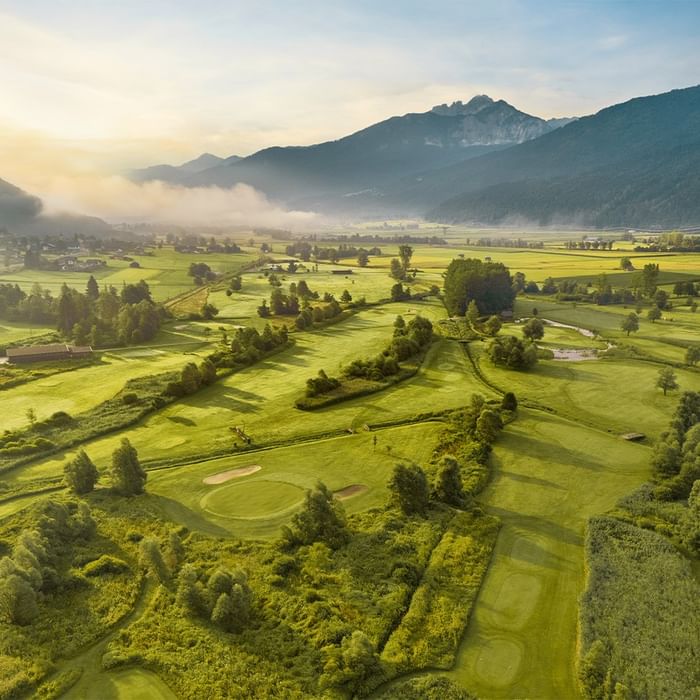 Luftaufnahme eines Golfplatzes mit grünen Feldern und Bergen im Hintergrund bei Falkensteiner Hotel & Spa Carinzia