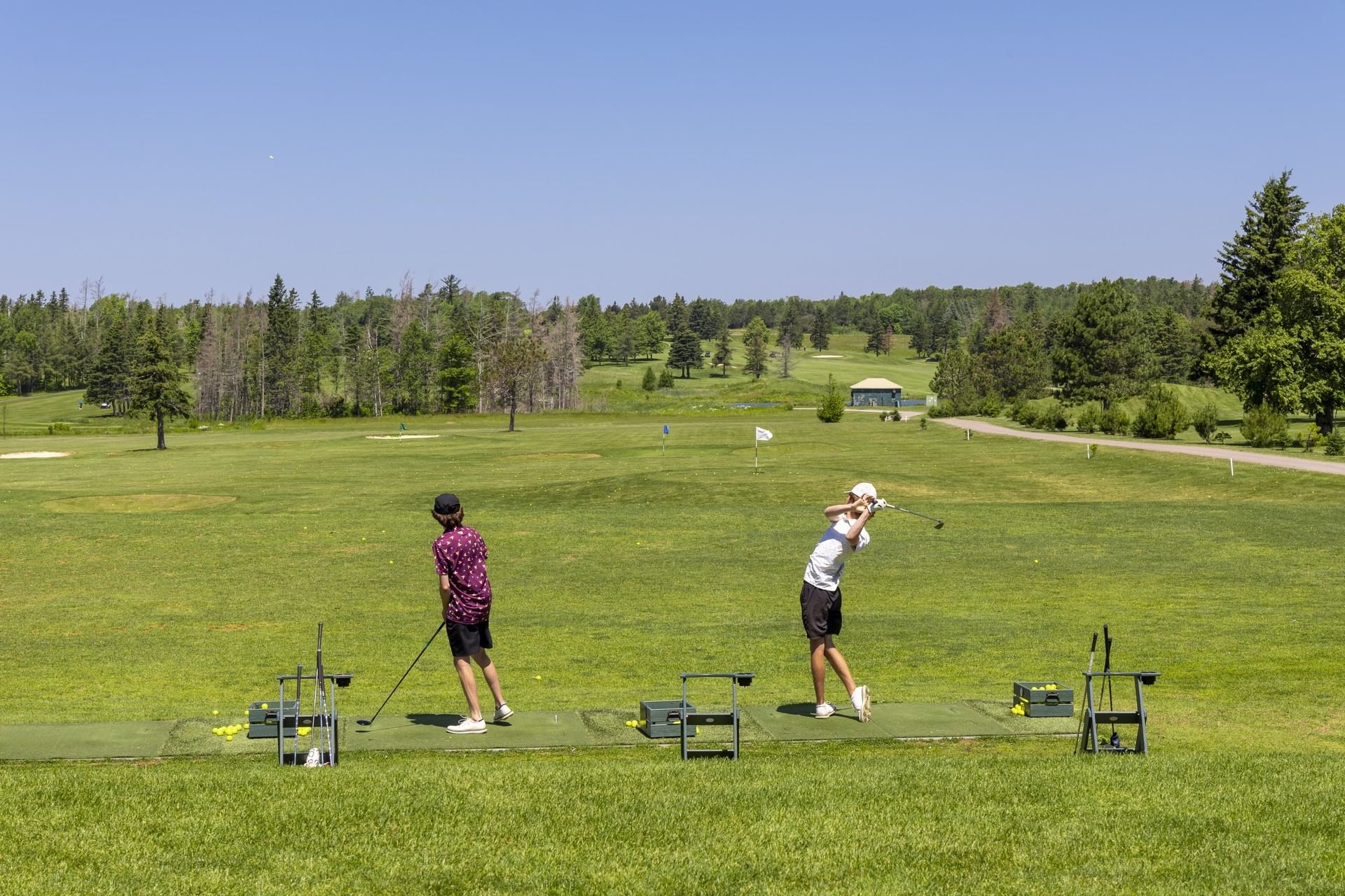 Golfers practicing swings on a range by green fields under a clear blue sky near Rodd Brudenell River Resort