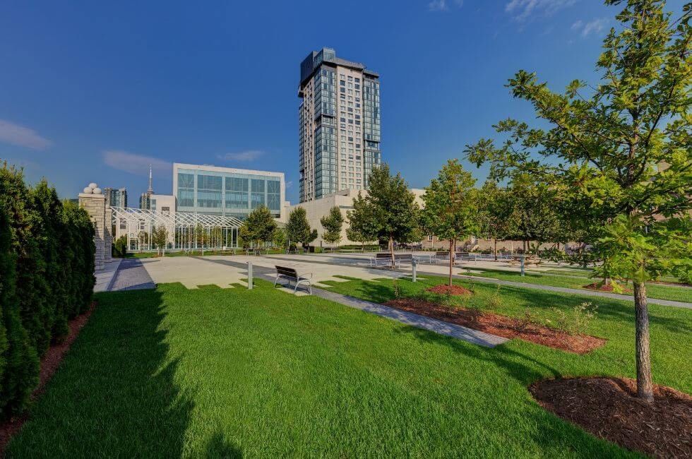 Hotel X Toronto tower rising by green lawns, benches and young trees under a clear blue sky in the landscaped grounds