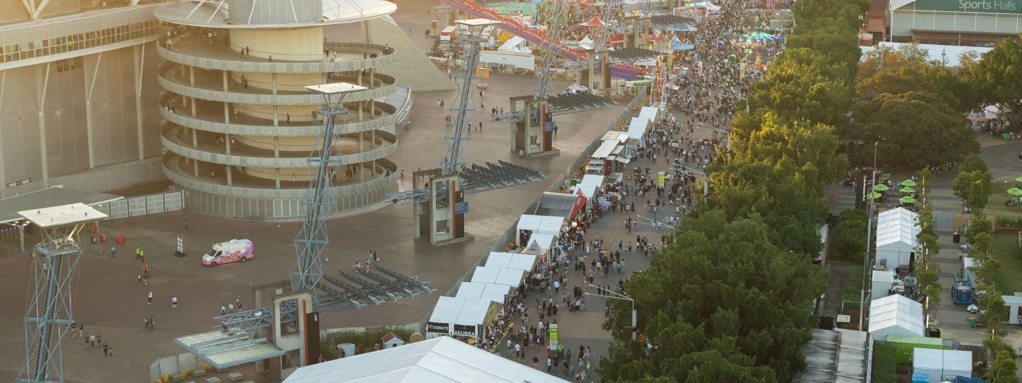 Aerial view of Sydney Showground with crowds, modern buildings, carnival rides, and green welcome tent at sunset