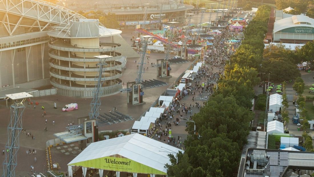 Aerial view of Sydney Showground with crowds, modern buildings, carnival rides, and green welcome tent at sunset
