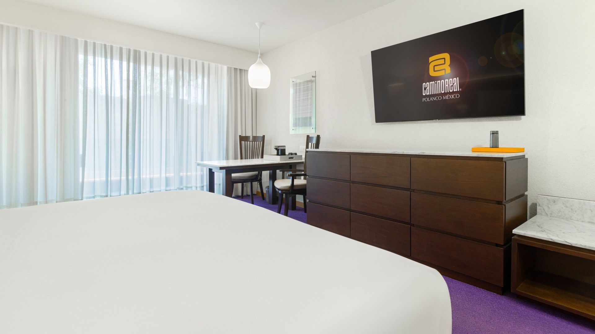 Habitación de hotel con cama, televisor, mesa y sillas, y ventana con cortinas.