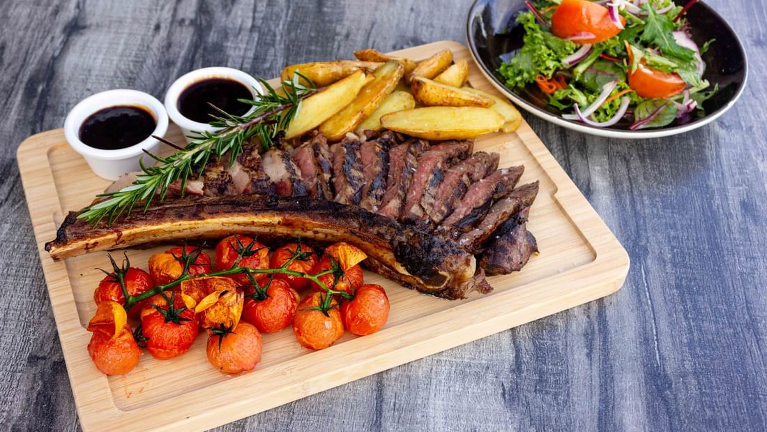 A wooden cutting board with steak, potatoes, roasted tomatoes, rosemary, salad, and two sauces on it.