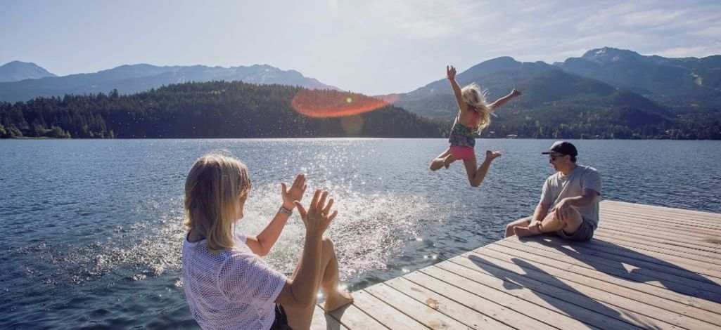 Family relaxing on a wooden dock at Whistler’s lakefront as a child jumps into the water with mountain views in the background.