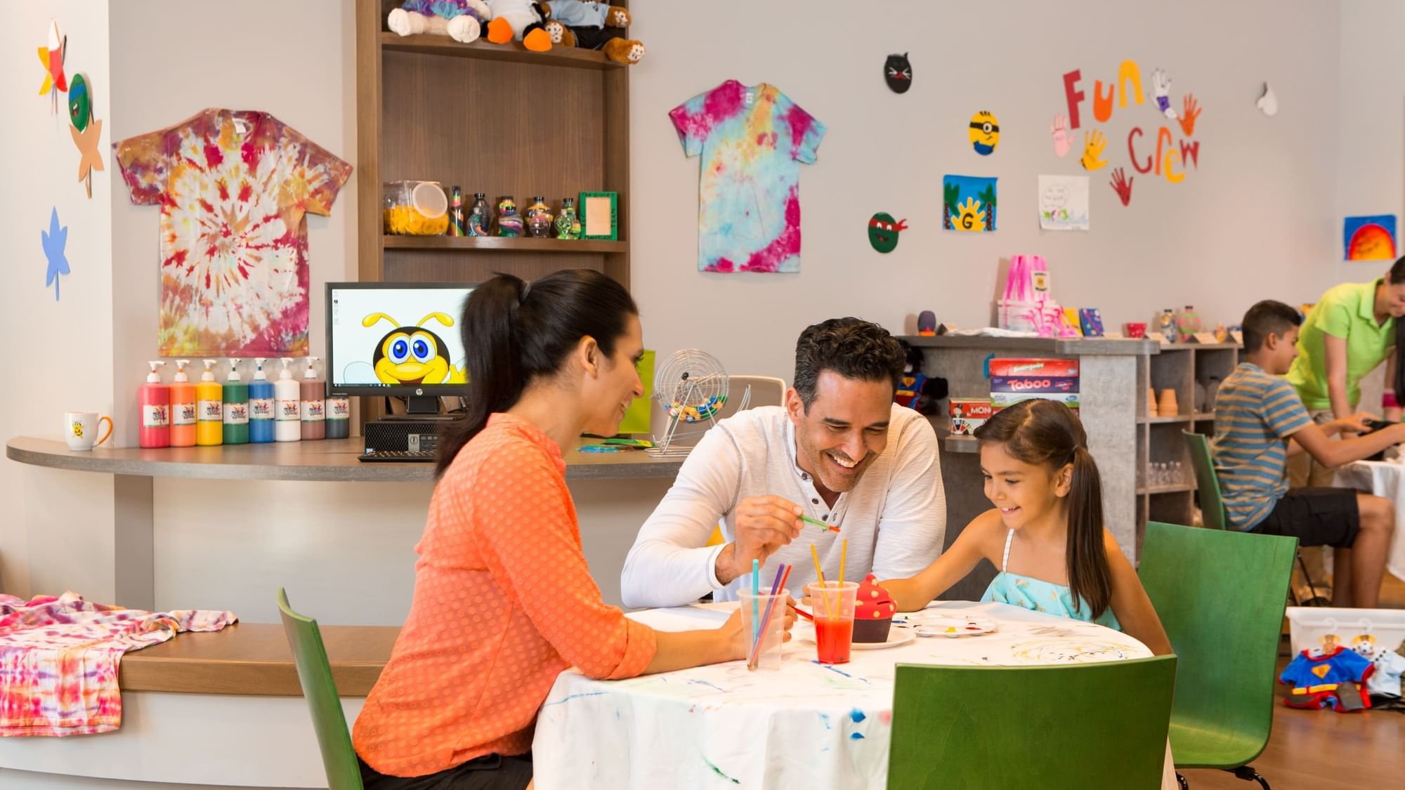 Family doing Father's Day crafts at a table in a brightly colored room with toys and decorations.