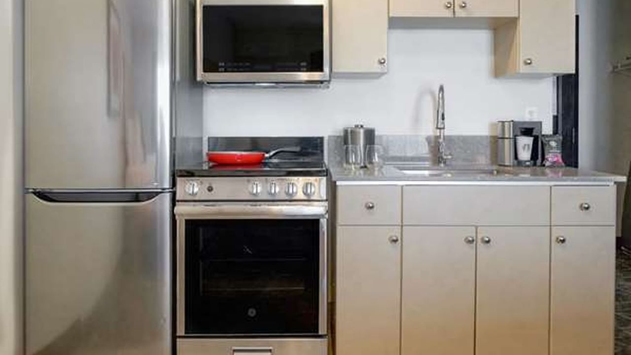 Compact kitchenette with stainless steel refrigerator, oven, microwave and sink, neatly arranged with beige cabinets.