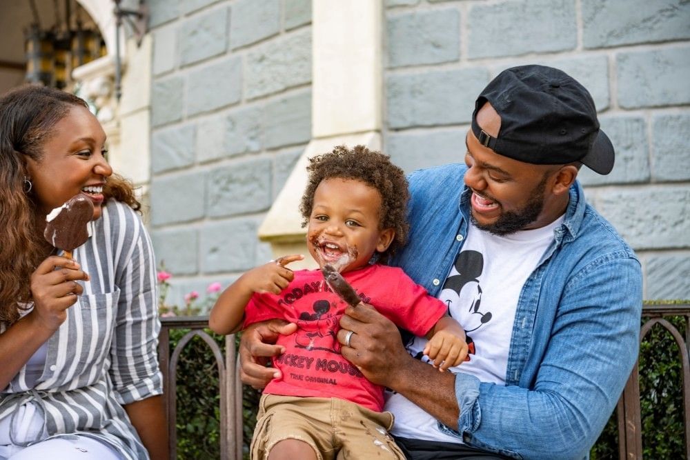 A toddler at Disney World in a red Mickey shirt sits between his parents as they enjoy Mickey ice cream bars, laughing.