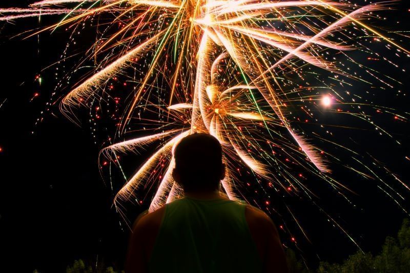 A silhouetted person looks up at a large burst of orange, yellow, green, and white fireworks in the dark night sky. 