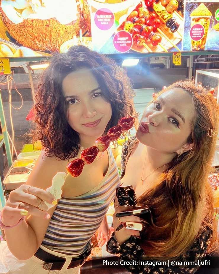 Close-up of two ladies enjoying tanghulu from a street food stall near Imperial Lexis Kuala Lumpur, enjoying Jalan Alor best food