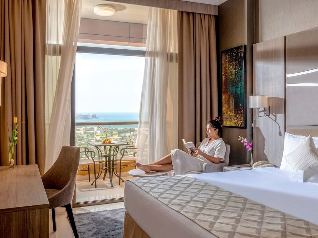 Modern hotel room with ocean view balcony, woman relaxing on chair with tablet, perfect for extended stays