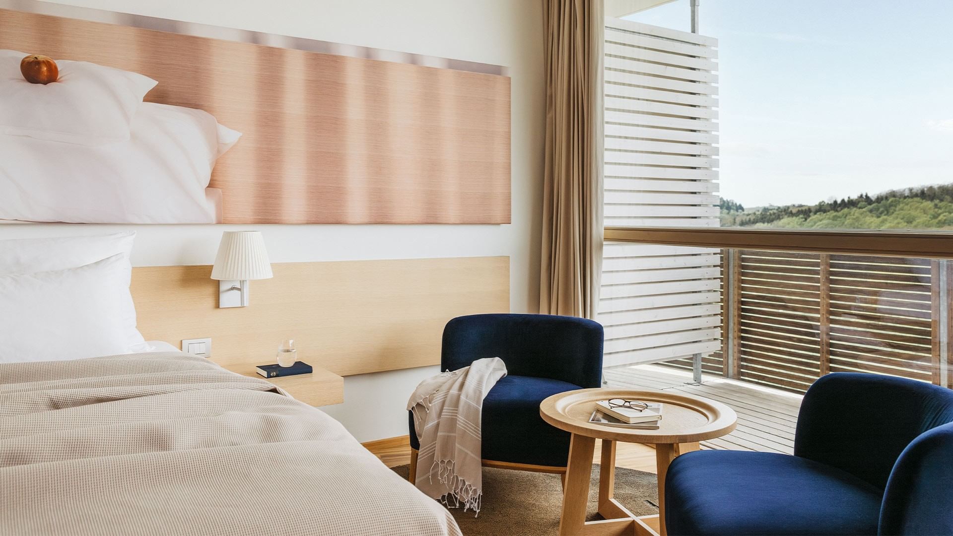 Modernes Hotelzimmer mit Bett, Leselampen, Tisch und zwei blauen Sesseln.