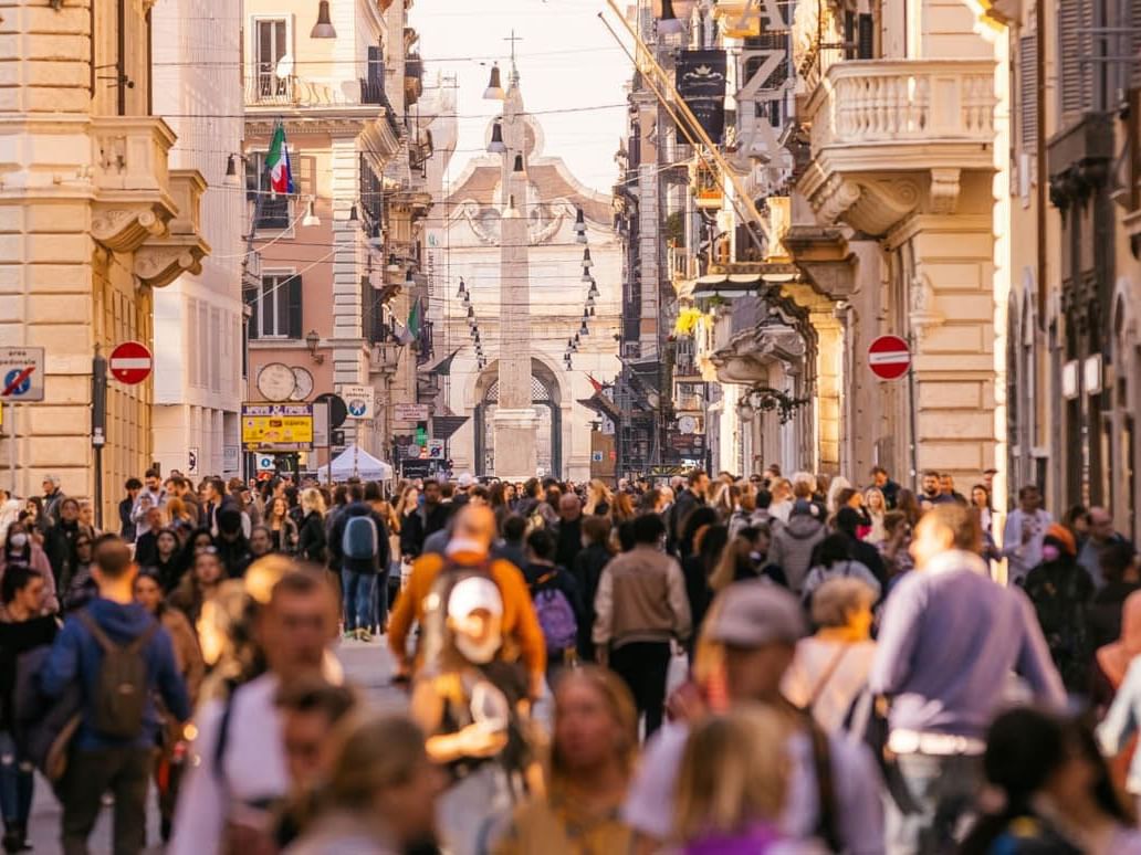Via del Corso, bustling with crowds, historic architecture, and shops, glowing in warm sunlight near The Guardian Hotel