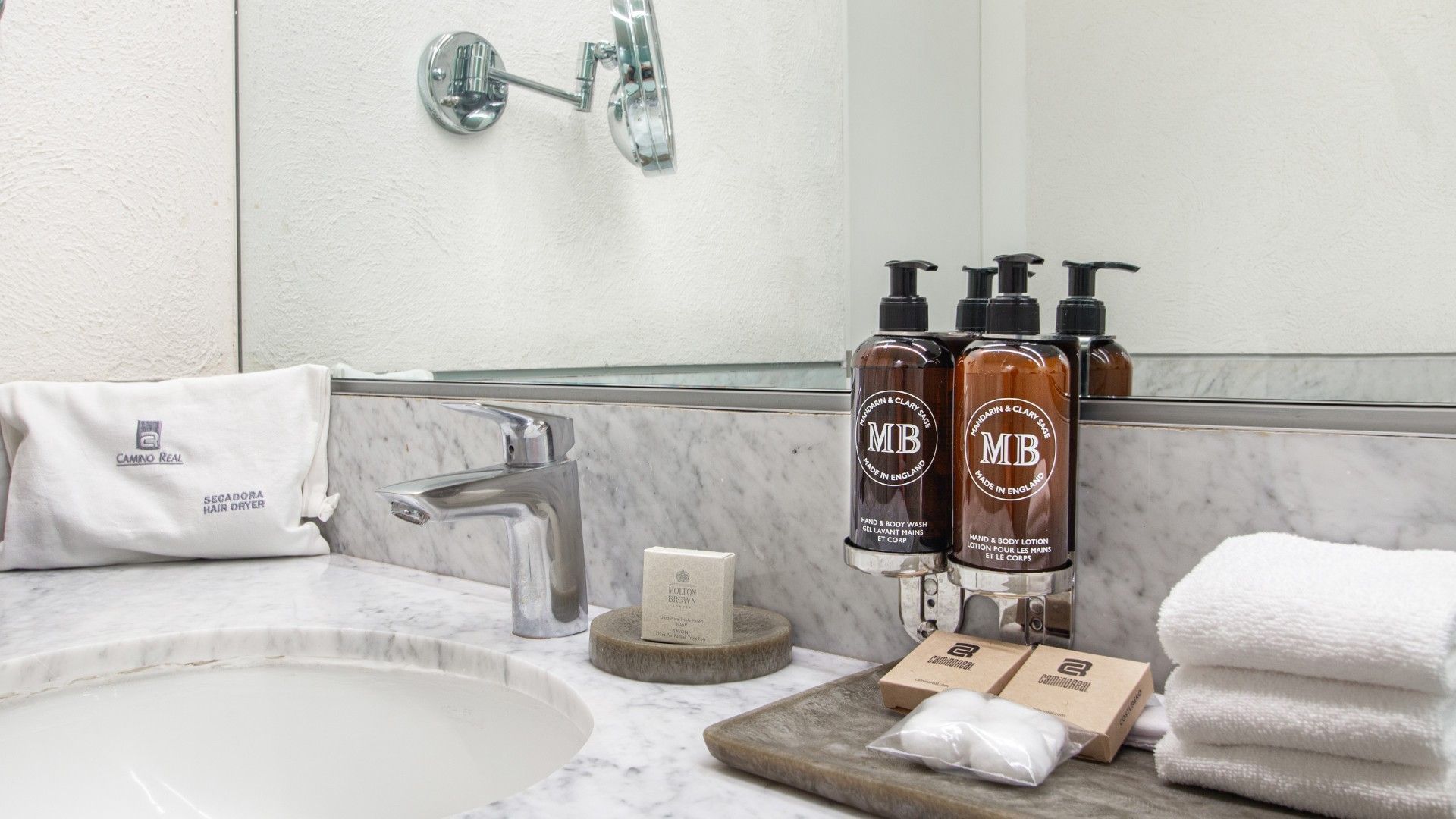 Baño con lavabo, espejo, toallas, cajas de jabón y tres botellas de MB.