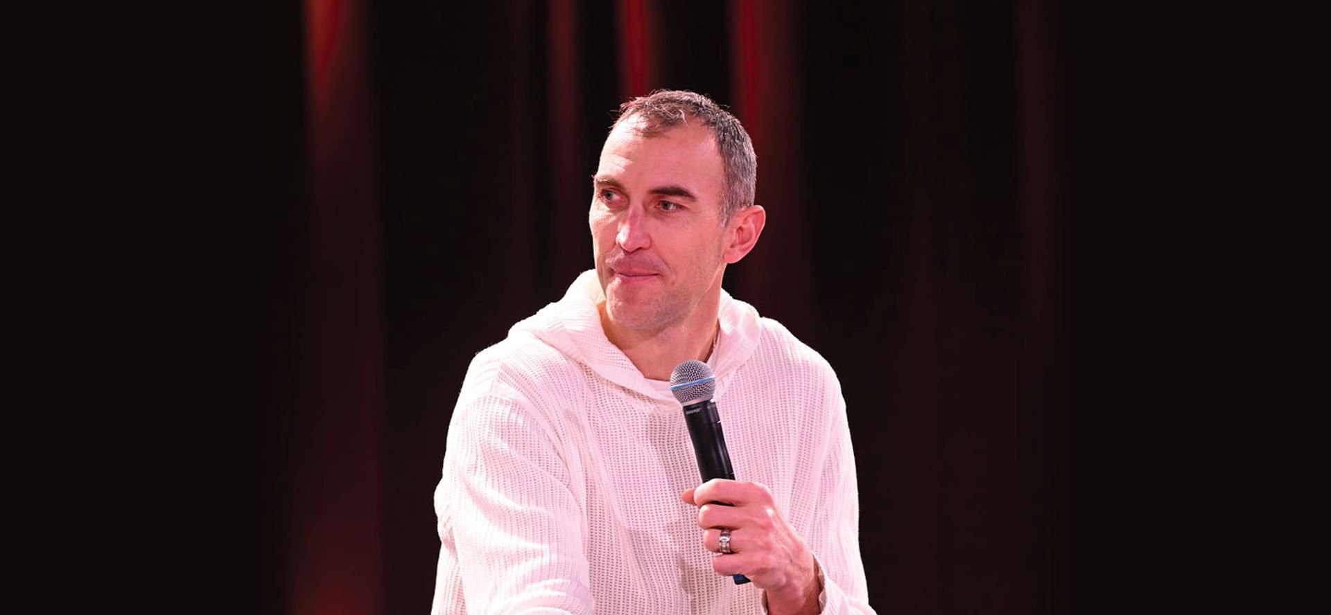 Comedian Mark Normand holding microphone on stage speaking to audience in front of backdrop at The Brook