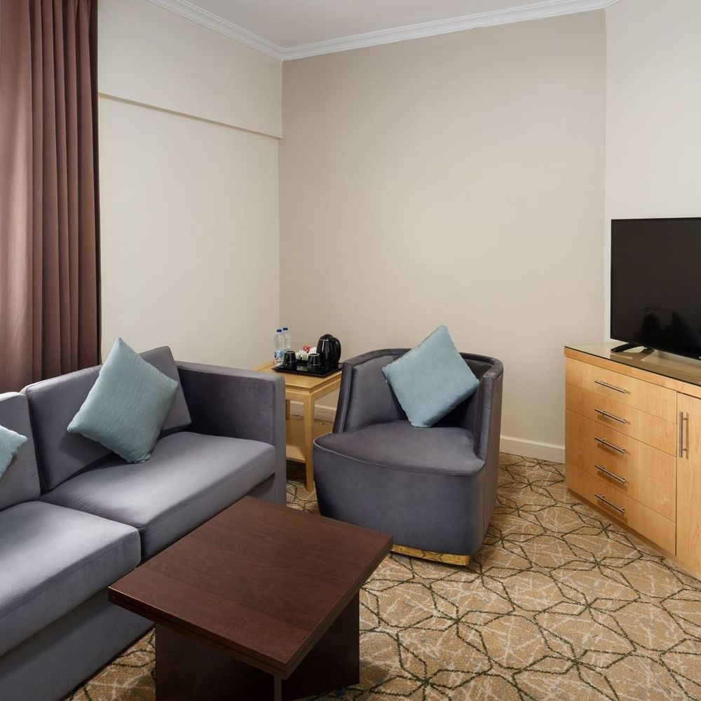 منطقة الجلوس في الجناح التنفيذي بفندق سجى ووريك المدينة، تضم أريكة رمادية مع وسائد ملوّنة، كرسيًا منفصلًا، طاولة قهوة خشبية، خزانة خشبية مع تلفاز بشاشة مسطحة، ومنضدة جانبية مزودة بخيارات الضيافة.