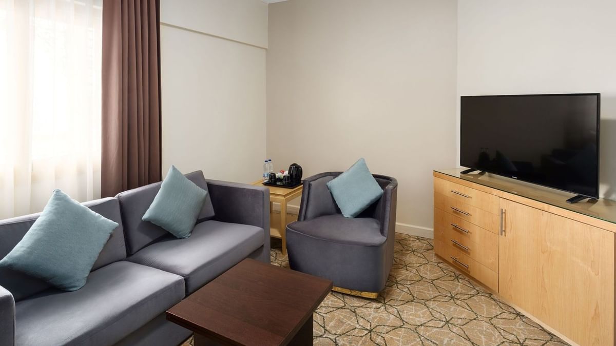 منطقة الجلوس في الجناح التنفيذي بفندق سجى ووريك المدينة، تضم أريكة رمادية مع وسائد ملوّنة، كرسيًا منفصلًا، طاولة قهوة خشبية، خزانة خشبية مع تلفاز بشاشة مسطحة، ومنضدة جانبية مزودة بخيارات الضيافة.