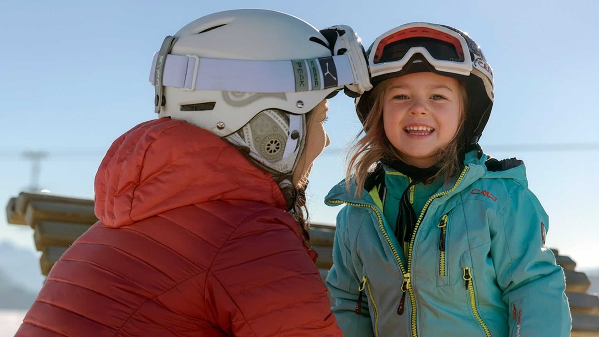 Una donna e una bambina sorridenti con caschi da sci in montagna presso Falkensteiner Group