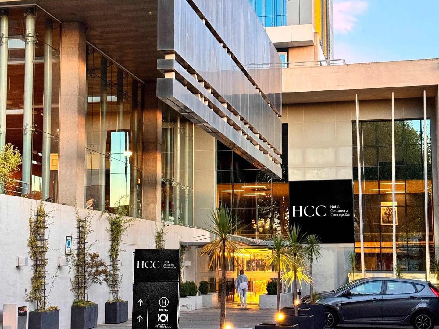 Entrada de un hotel moderno con letreros negros, fachada acristalada y un coche aparcado en la puerta.