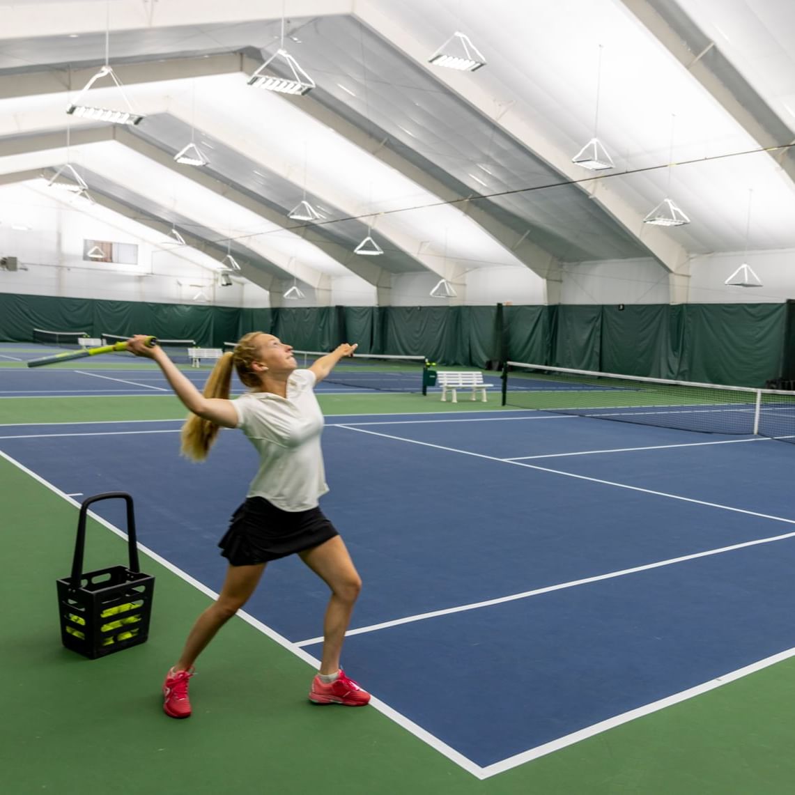 Tennis player in action on a blue court with a ball basket nearby.