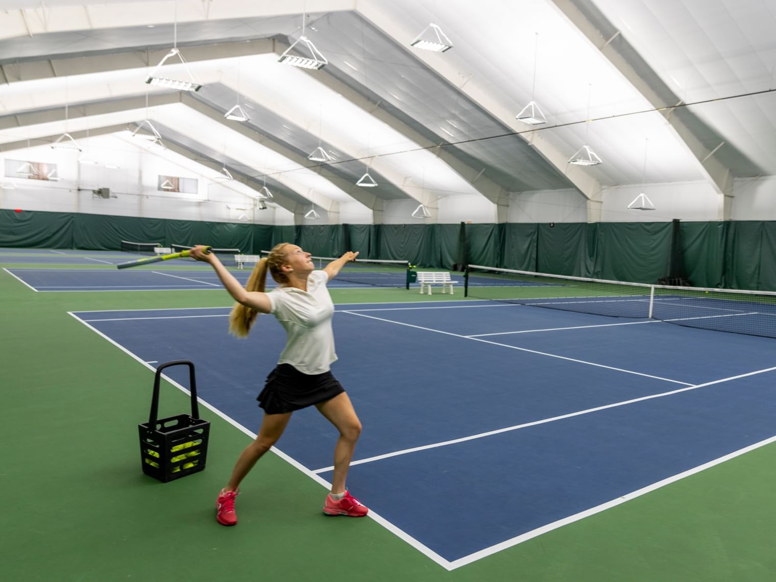 Tennis player in action on a blue court with a ball basket nearby.