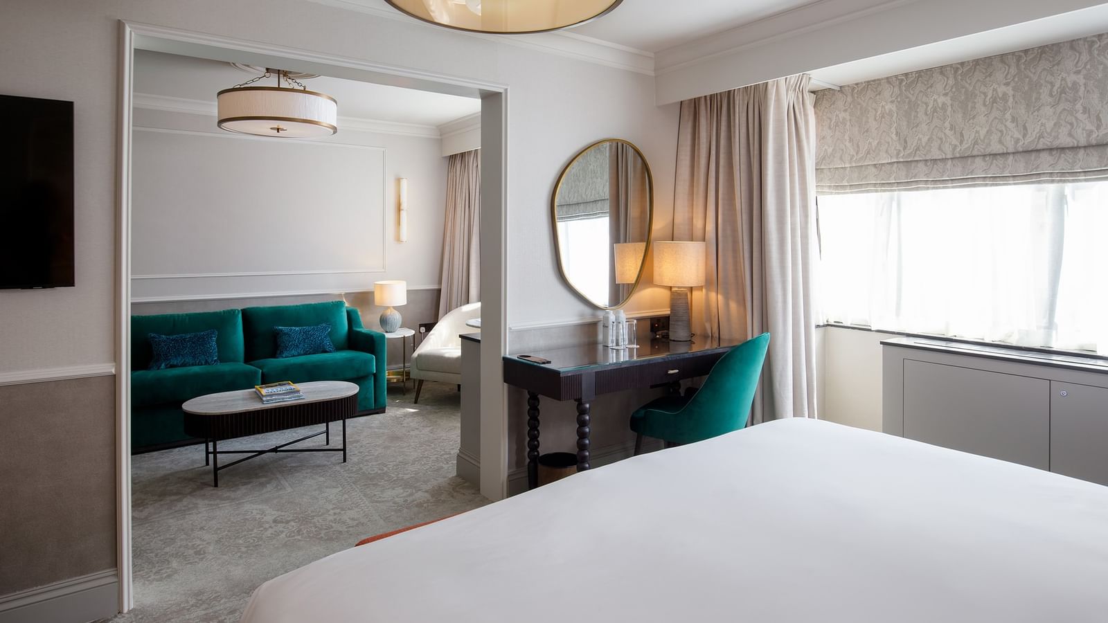 Suite élégante avec lit king-size, bureau en bois, miroir rond et salon adjacent avec canapé turquoise et table basse.