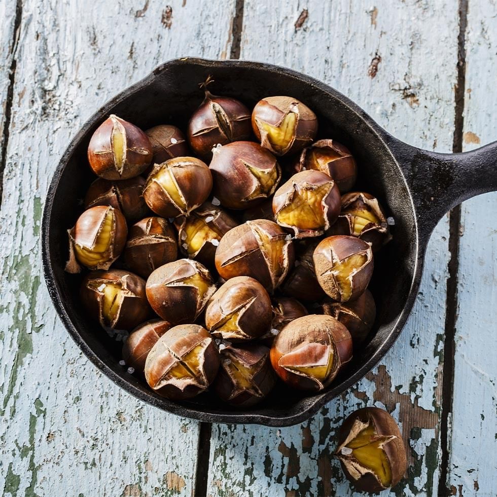 Roasted chestnuts in a cast iron skillet on a blue wooden surface with one chestnut beside the skillet.