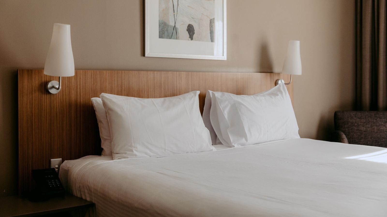 A hotel room with a neatly made bed, two white pillows, and framed artwork above it.